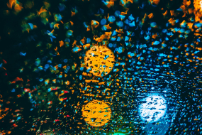 Eine Nahaufnahme von Regentropfen auf einer Fensterscheibe bei Nacht, die bunte Lichter von Straßenlaternen und Fahrzeugen im Hintergrund reflektieren. Die Lichter erscheinen verschwommen und in leuchtenden Farben wie Gelb, Blau, Rot und Grün, die durch die Tropfen in bokehartigen Mustern verzerrt werden. Das Bild vermittelt eine atmosphärische, abstrakte Stimmung mit einem starken Fokus auf Licht- und Farbkontraste.