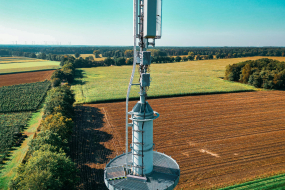 Die Fotografie zeigt einen Mobilfunkmast in einer ländlichen Umgebung. Der Mast steht zentral im Bild und ist mit mehreren Antennen und technischen Geräten ausgestattet. Im Hintergrund erstreckt sich eine weite, landwirtschaftlich genutzte Fläche mit verschiedenen Feldern, die von Baumreihen begrenzt sind. Der Himmel ist klar und türkisblau, was auf sonniges Wetter hinweist. Am Horizont sind Windkraftanlagen erkennbar, die in die Landschaft eingebettet sind.