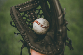 Eine Hand hält einen schwarzen Baseballhandschuh mit sichtbaren Nähten und Netzstruktur in der Mitte. Im Handschuh liegt ein leicht abgenutzter Baseball mit roten Nähten. Der Hintergrund ist unscharf und zeigt eine grüne Rasenfläche, vermutlich ein Spielfeld. Die Perspektive ist aus der Sicht der Person, die den Handschuh trägt, mit Fokus auf Ball und Handschuh.