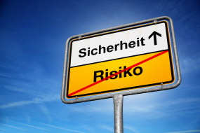 Ein gelb-weißes Straßenschild mit schwarzer Schrift steht vor einem strahlend blauen Himmel. Das obere, weiße Feld zeigt das Wort „Sicherheit“ mit einem nach oben zeigenden Pfeil. Das untere, gelbe Feld enthält das Wort „Risiko“, das mit einem roten diagonalen Strich durchgestrichen ist. Das Schild erinnert an Ortsschilder und symbolisiert die Bewegung von Risiko hin zu mehr Sicherheit.