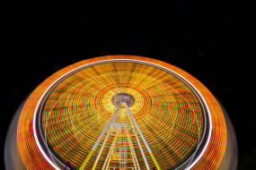 Ein großes Riesenrad dreht sich in der Dunkelheit der Nacht. Durch die Langzeitbelichtung entsteht ein faszinierender Lichteffekt: Die bunten LED-Leuchten des Riesenrads formen konzentrische Kreise in Gelb-, Rot-, Grün- und Weißtönen. Die Konstruktion des Riesenrads mit gelben und weißen Stützen ist in der unteren Bildhälfte zu sehen, während der dunkle Nachthimmel den leuchtenden Effekt verstärkt.