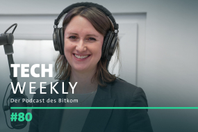 Bitkom-Politik-Expertin Sophie Vogt-Hohenlinde mit schulterlangem braunem Haar steht im Podcast-Studio und lächelt in die Kamera. Sie trägt große schwarze Over-Ear-Kopfhörer und ein dunkelgraues Blazer-Jackett über einem grauen Oberteil. Vor ihr steht ein professionelles Mikrofon mit schwarzem Schwenkarm. Der Hintergrund ist ein neutraler, heller Raum mit einer geschlossenen Tür. Überlagert auf dem Bild steht der Titel des Podcasts: „TECH WEEKLY – Der Podcast des Bitkom“, sowie die Episodennummer „#80“.