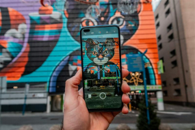 Eine Hand hält ein Smartphone vor einem großen Wandgemälde, das eine bunte, geometrisch gestaltete Katze zeigt. Das Smartphone zeigt die Kamera-App mit dem Live-Bild der Fassade, wobei die Katze auf dem Display perfekt mit der Wand dahinter übereinstimmt. Das Wandgemälde bedeckt die gesamte Seite eines Gebäudes und besteht aus leuchtenden Farben wie Blau, Orange und Schwarz. Die Szene spielt sich in einer städtischen Umgebung mit weiteren Gebäuden im Hintergrund ab.