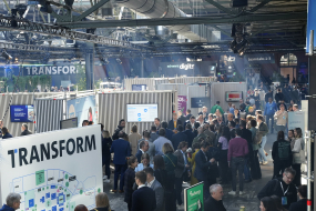 Die STATION Berlin ist gefüllt mit zahlreichen Menschen, die sich in Gruppen unterhalten oder durch die Gänge zwischen den Ständen bewegen. Im Vordergrund steht eine große Infotafel mit der Aufschrift „TRANSFORM“ und einer Übersichtskarte der Veranstaltung. Mehrere Personen betrachten diese Tafel. Im Hintergrund befinden sich zahlreiche Messestände mit Bildschirmen und Bannern, während Besucher geschäftig netzwerken. Die Halle ist industriell gestaltet mit hohen Decken und Stahlträgern.