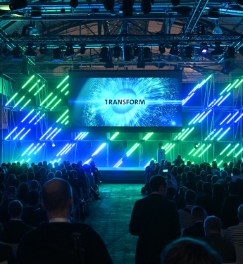 Publikum in dunkler Business-Kleidung sieht in einem Saal zu einer Bühne auf, wo ein Banner mit dem Wort "TRANSFORM" und einem sternähnlichen visuellen Effekt präsentiert wird. Die Szene ist in blaues und grünes Licht getaucht.