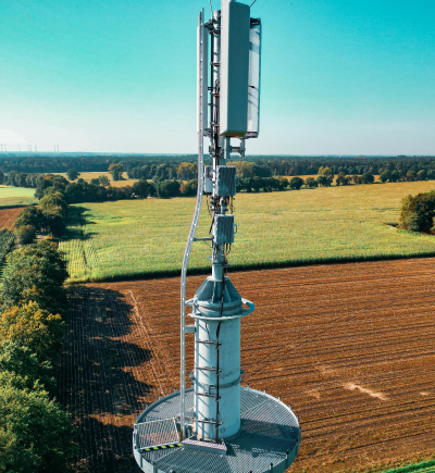 Die Fotografie zeigt einen Mobilfunkmast in einer ländlichen Umgebung. Der Mast steht zentral im Bild und ist mit mehreren Antennen und technischen Geräten ausgestattet. Im Hintergrund erstreckt sich eine weite, landwirtschaftlich genutzte Fläche mit verschiedenen Feldern, die von Baumreihen begrenzt sind. Der Himmel ist klar und türkisblau, was auf sonniges Wetter hinweist. Am Horizont sind Windkraftanlagen erkennbar, die in die Landschaft eingebettet sind.